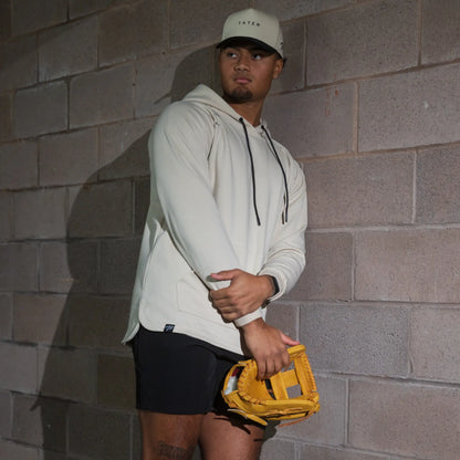 In the photo, a baseball player is wearing a cream-colored TATER-branded hoodie and cap, with a yellow infielder's glove, showcasing athletic wear suitable for both sports and leisure.