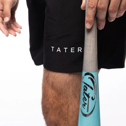 The image features a close-up view of a person wearing black athletic shorts with the word "TATER" printed on them, indicating they are part of a sports or athletic wear collection. The individual is also holding a light blue baseball bat, which is partially visible in the shot. The focus on the branded shorts and the bat suggests that the image might be used for promotional purposes to highlight the brand's association with baseball and athletic gear.