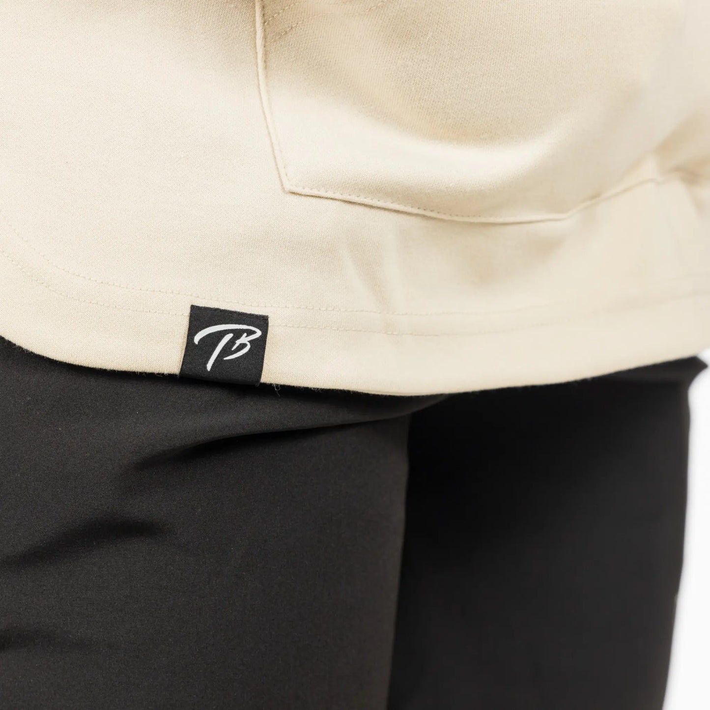 The image features a close-up view of a cream-colored hoodie with a small, square black logo label that has a white "TB" inscription, representing the brand identity. The hoodie fabric appears to be of a thick, durable quality suitable for athletic wear.