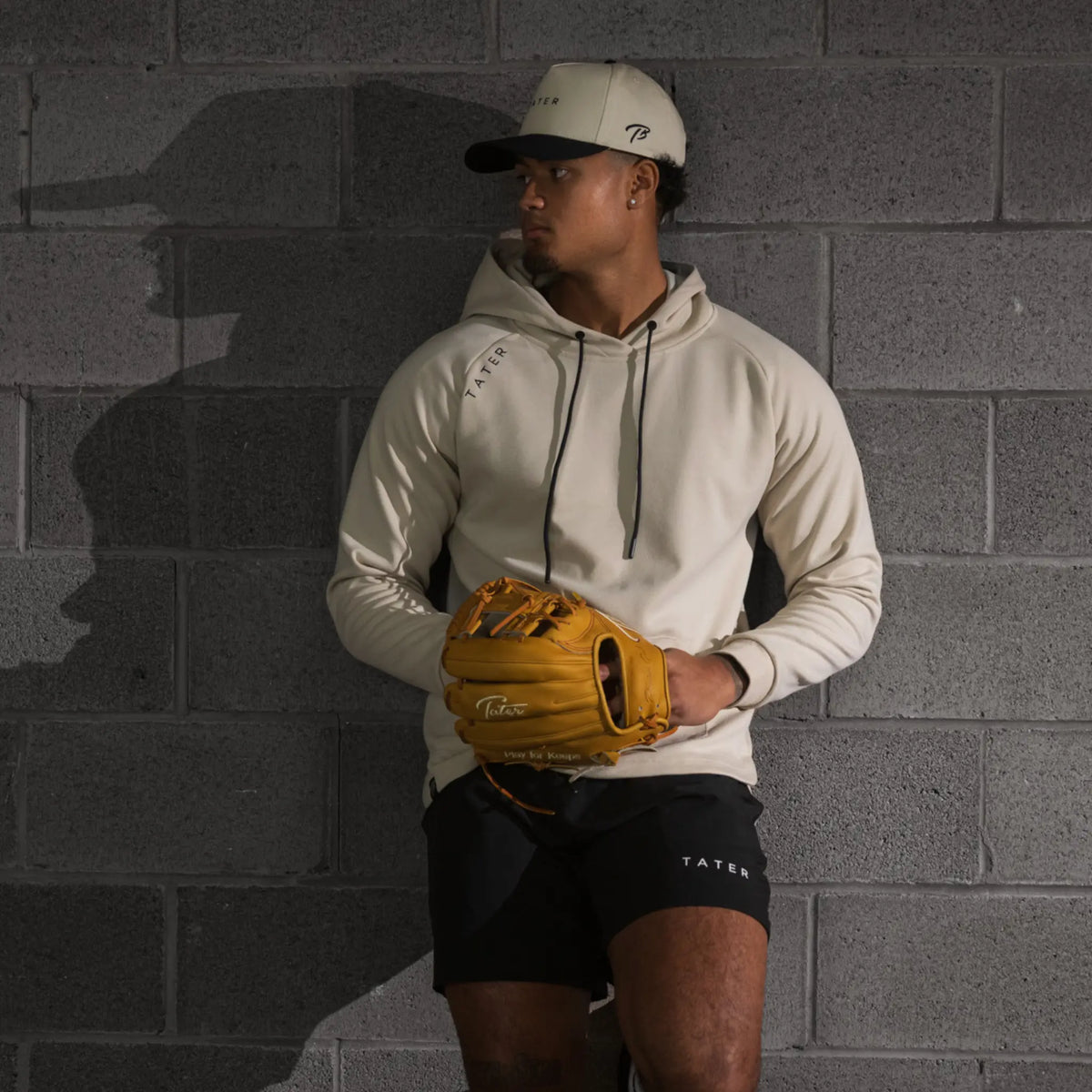 The uploaded image shows a college baseball player posed against a textured wall, capturing a strong athletic stance. The player is outfitted in TATER baseball brand gear, wearing a cream-colored hoodie with the TATER logo prominently displayed on the side, black athletic shorts also bearing the TATER logo, and a baseball cap with the same branding. He is holding a golden yellow baseball glove, adding a vibrant contrast to the neutral tones of the apparel. 