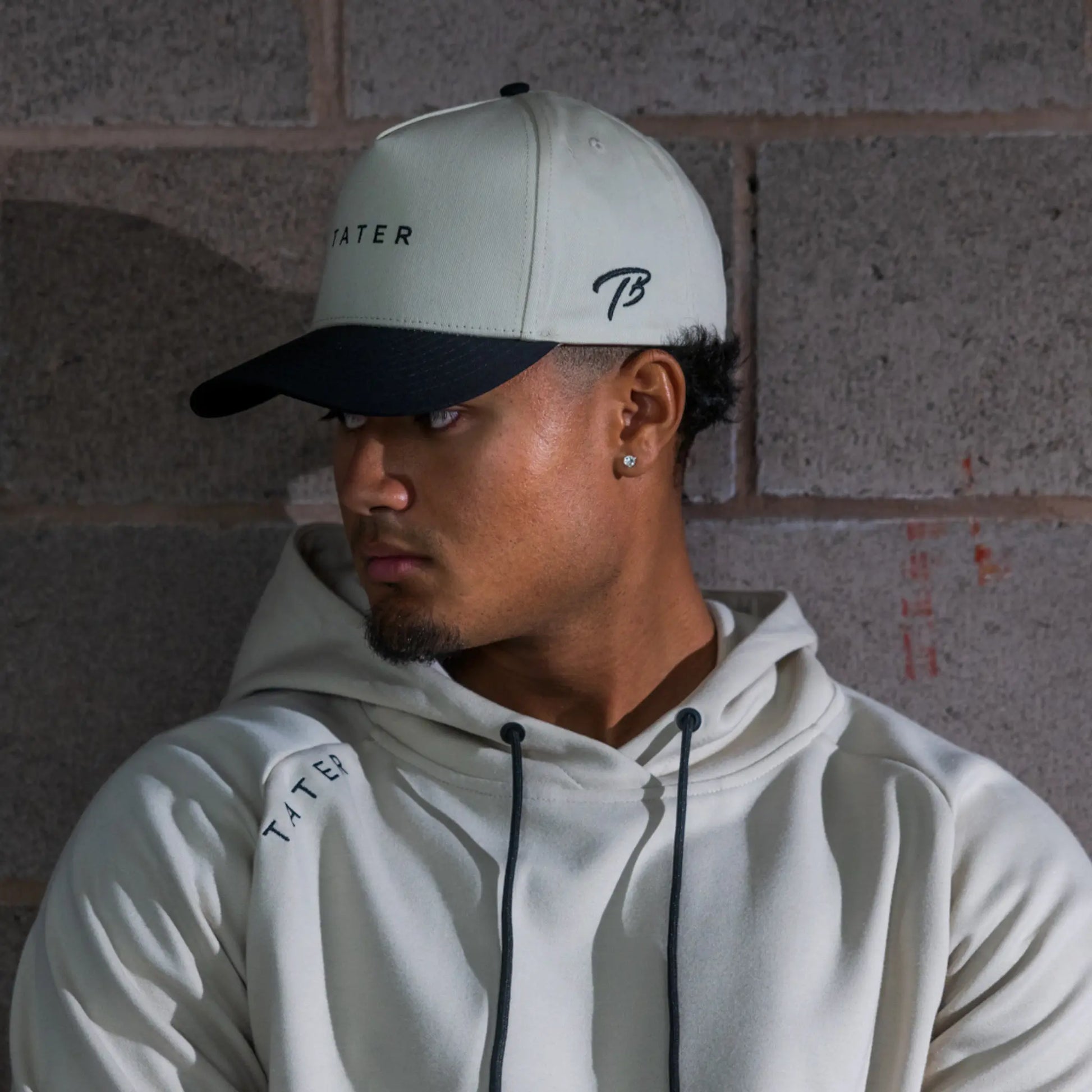 In this image, the subject is wearing a cream-colored snapback hat with the "Tater" logo in black, which matches the hoodie's color and branding. The hat's bill is black, creating a stylish contrast. The person's sideways glance adds a contemplative or focused ambiance to the photo. The lighting accentuates the textures and colors of the apparel, and the background's muted tones allow the Tater Baseball brand elements to stand out, emphasizing a fusion of casual style and athletic wear.