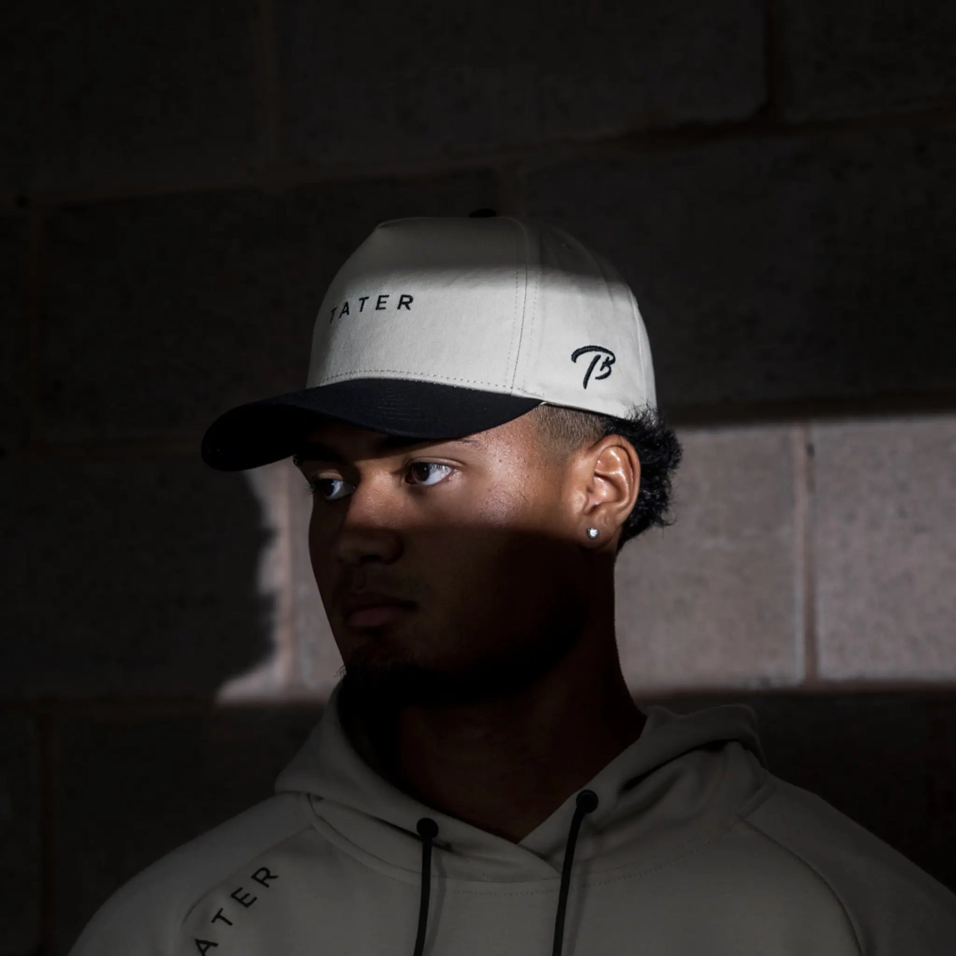 The image features a person wearing a tan snapback hat with the word "TATER" on the front and the Tater Baseball "TB" logo on the side, paired with a hoodie that also carries the "TATER" branding. The overall look suggests a coordinated athletic style, with the person looking off to the side, creating a contemplative and focused atmosphere. The lighting in the photo highlights the hat and the person's profile, adding to the moody and professional aesthetic of the apparel.