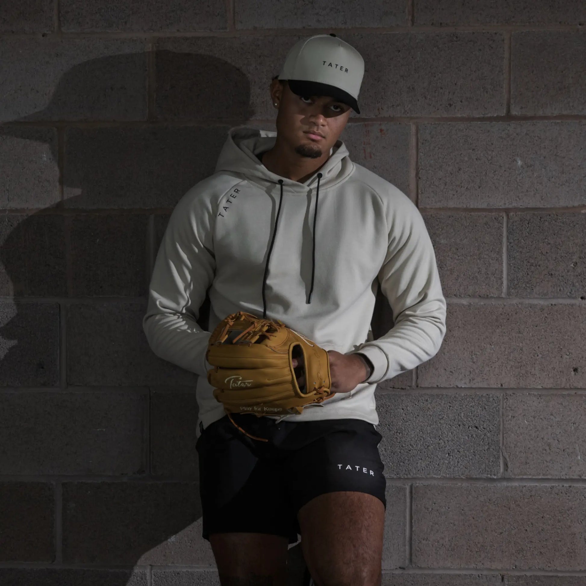 The image showcases an individual donning a coordinated sports ensemble from Tater Baseball. The person is wearing a cream snapback hat with the brand's name and logo, complemented by a matching hoodie. In one hand, they hold a gold-colored baseball glove, and they're dressed in black athletic shorts bearing the brand's name. The overall styling presents a sleek, athletic look, with a backdrop that adds a raw, urban feel to the image, highlighting the sporty yet stylish nature of the apparel. 