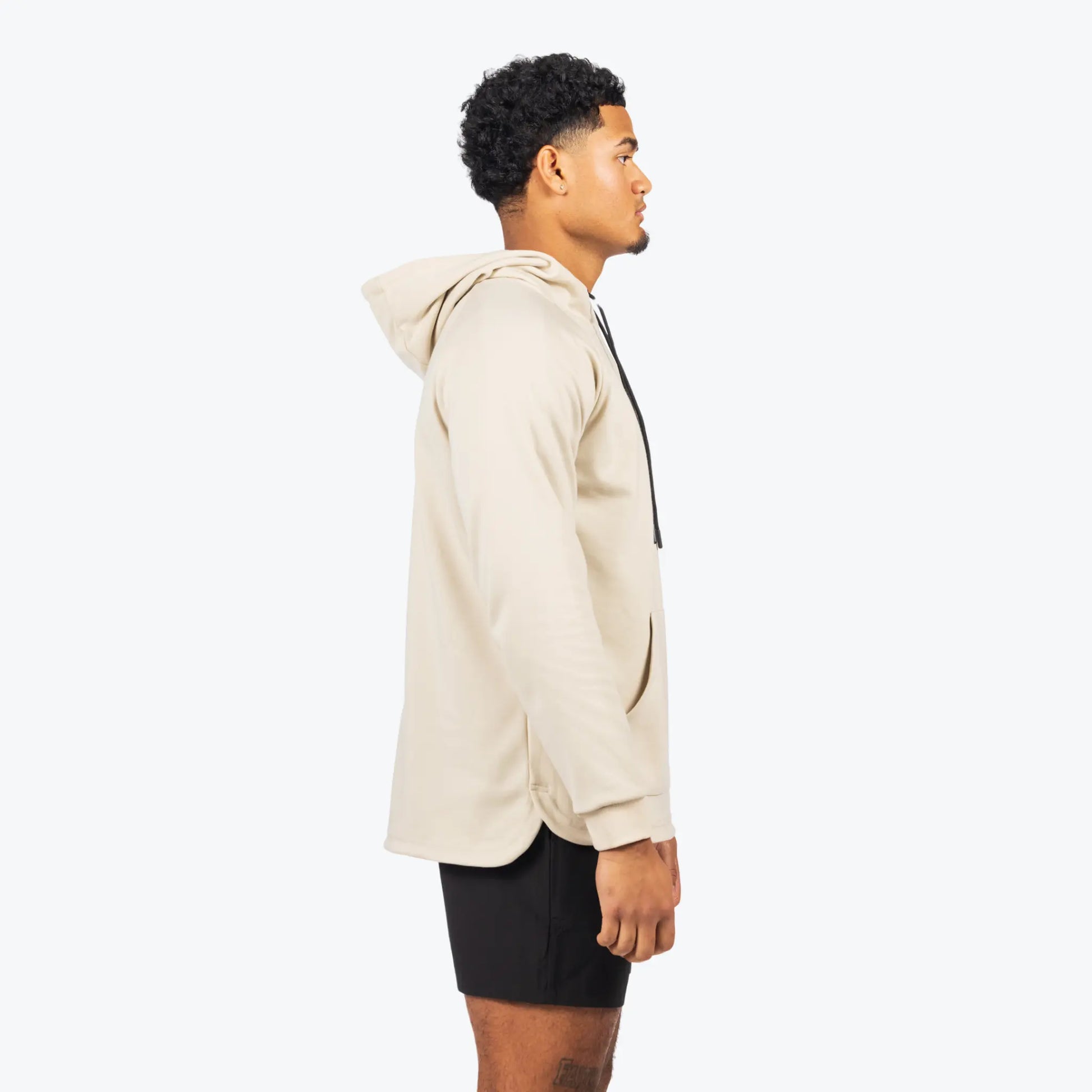 The uploaded image features a side view of a model wearing a professional baseball training hoodie with long sleeves. The hoodie is cream-colored, with a drawstring hood and the "TATER" logo printed along the sleeve, pairing well with the black athletic shorts the model is wearing. The attire highlights a combination of comfort, style, and functionality, suitable for both training and casual wear within a baseball lifestyle context.