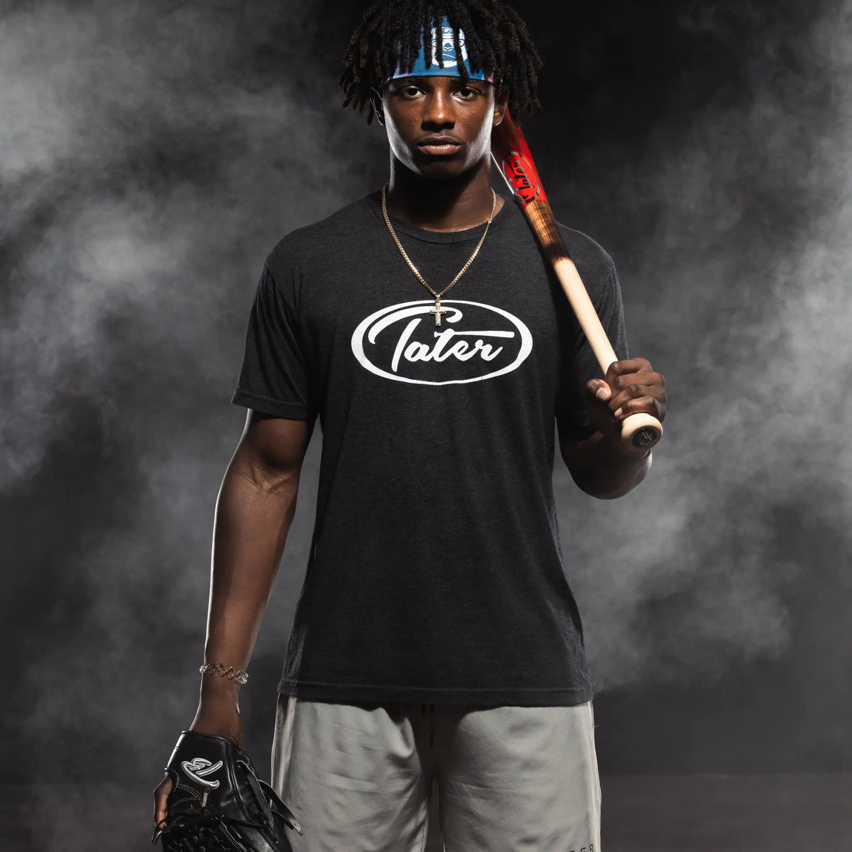 The image features an athlete clad in Tater Baseball gear, with a black logo tee, grey shorts, a baseball bat, and a glove, set against a dramatic smoky backdrop for a promotional look.