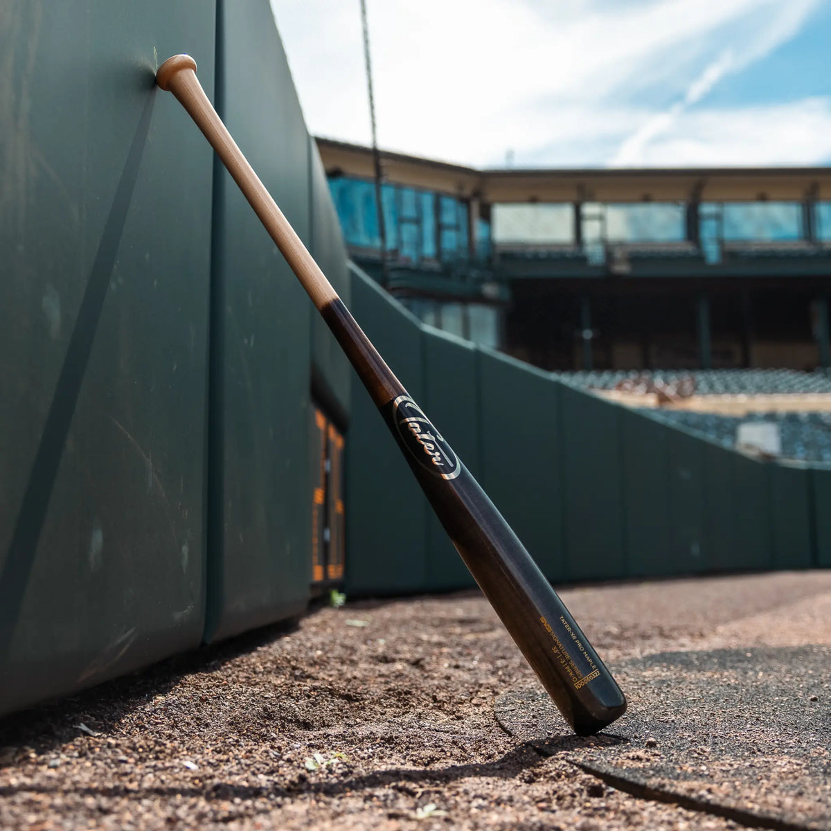 Premium maple wood baseball bat model X6 with a slight end-load, 32-33 inch, drop 3, resting against a dugout wall in a professional baseball stadium.