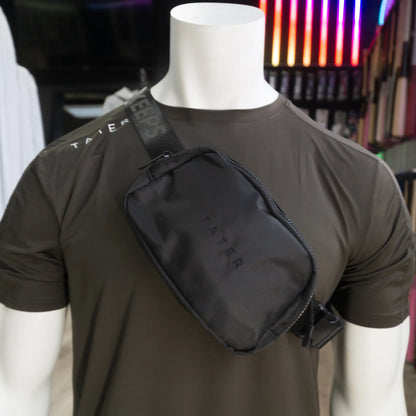 The image presents a mannequin dressed in an olive athletic T-shirt with a black crossbody fanny pack slung over the shoulder. The fanny pack features the Tater brand name embossed on the front, while the strap is adorned with the phrase "KEEPERS" in a repeating pattern, contributing to the sporty and stylish aesthetic of the product. The combination of the crossbody pack with the athletic wear suggests a trendy and practical accessory for sports enthusiasts or individuals with an active lifestyle. 