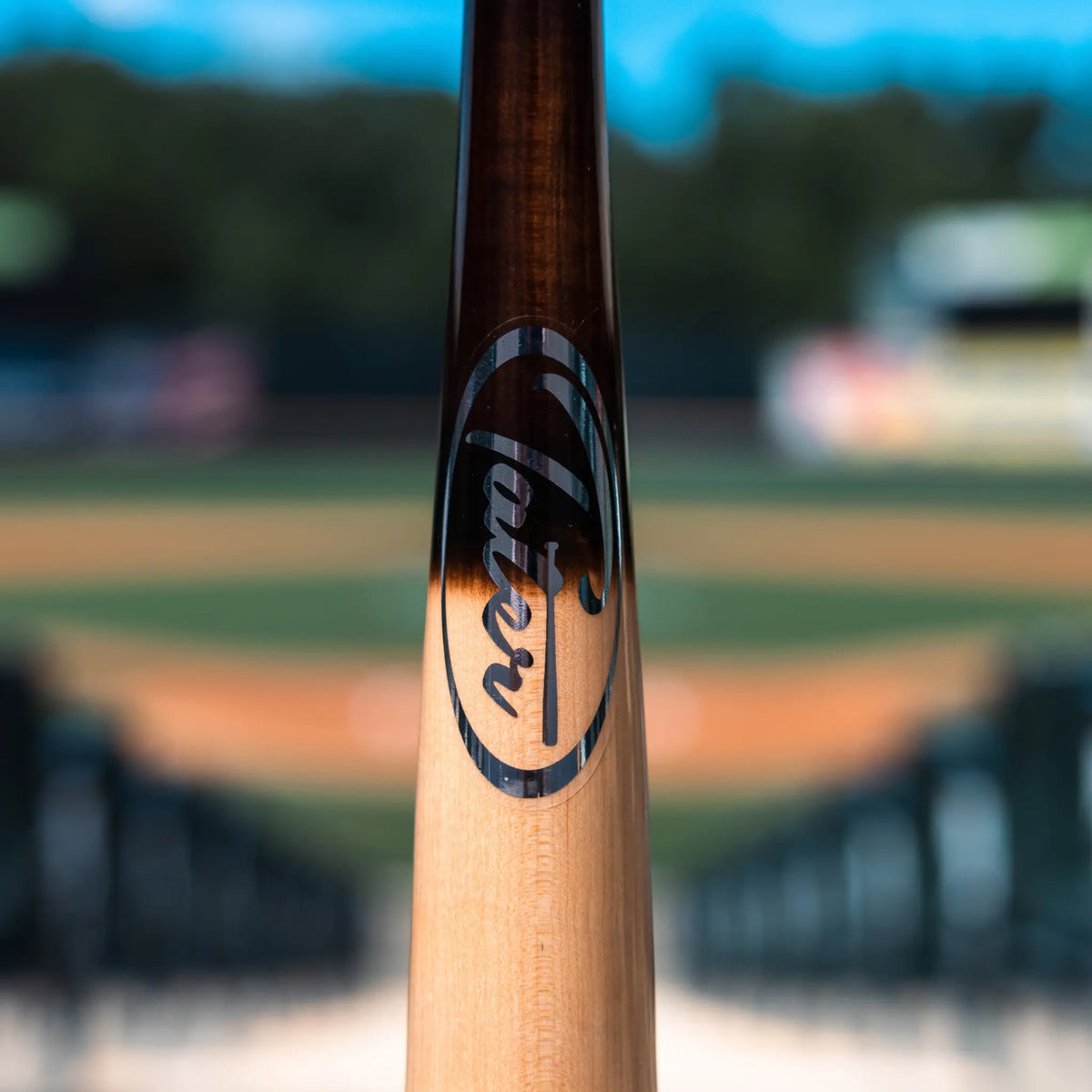 Featured in this image is the middle section of a Tater X12 Pro Maple baseball bat, displaying the elegant sliver Tater logo on its polished wood surface. The bat is centered against a softly focused baseball diamond, capturing the essence of the game.