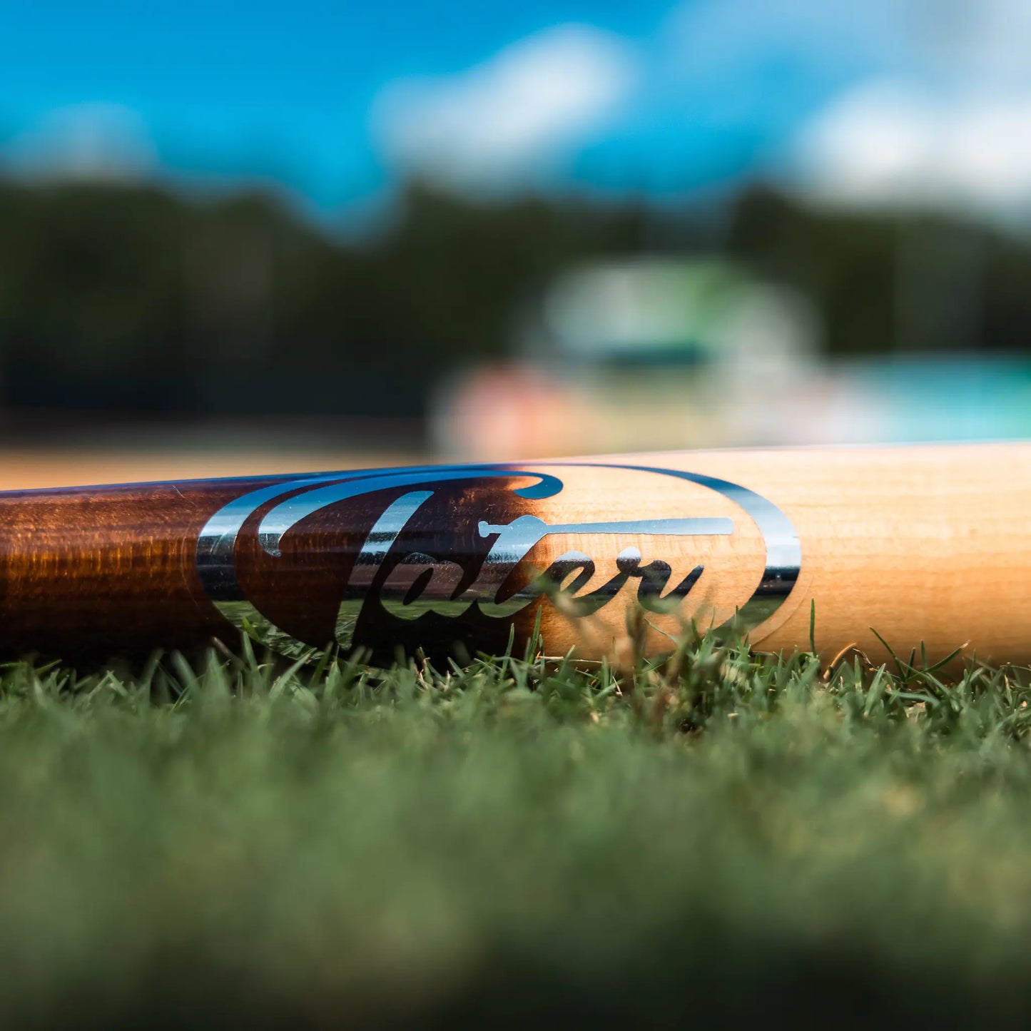 An image showcasing the popular Tater X12 Pro Maple bat, which stands out for its performance on the field, sturdy construction, and timeless style that appeals to competitive players. Bat is resting on grass, closeup of Tater logo.This image captures a close-up view of a Tater X12 Pro Maple bat lying horizontally on the grass. The focus is on the bat's barrel which features the bold, black Tater logo, with a blurred baseball diamond in the background under a bright, clear sky.
